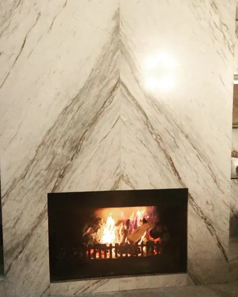 Stone with mirrored veining is used for the fireplace surround, alongside comfortable seating and a restrained neutral palette. The veining forms a sharp symmetrical arrangement above the fire.