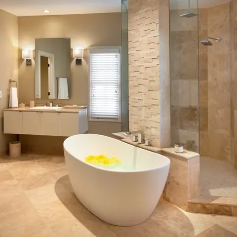 Warm travertine is used for the bathroom walls and floor, alongside a freestanding white bath and soft lighting.